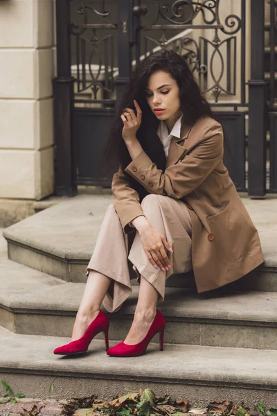Uzun, esmer, kıvırcık saçlı, binanın arka planında poz veren güzel bir kadının portresi. Merdivenlerde oturuyor. parlak kırmızı yüksek topuklu ayakkabılar — Stok fotoğraf