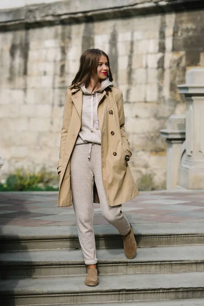 Portrait of fashionable women in beige sports suit, trench coat and stylish suede loafer posing on the stone stairs — Stock Photo, Image