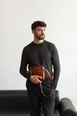 Koyu gri takım yakalı şık bir adam konyak deri bir çanta ile minimalist beyaz stüdyo arka planına karşı poz veriyor.