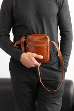 Beyaz bir stüdyo arka planına karşı konyak derisi dikey kurye çantası giyen, yaka yaka yakalı bir erkek model ve özel dikey pantolon giyiyor.