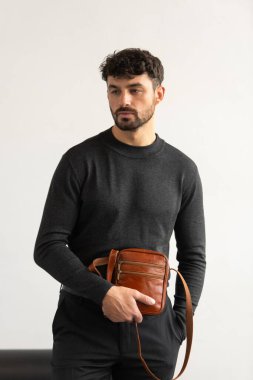 Koyu gri takım yakalı şık bir adam konyak deri bir çanta ile minimalist beyaz stüdyo arka planına karşı poz veriyor.
