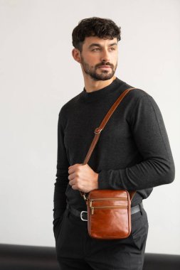 Koyu gri takım yakalı şık bir adam konyak deri bir çanta ile minimalist beyaz stüdyo arka planına karşı poz veriyor.