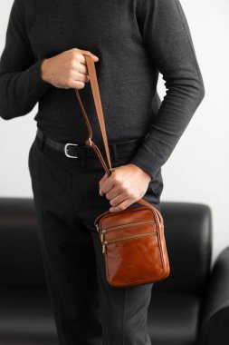 Beyaz bir stüdyo arka planına karşı konyak derisi dikey kurye çantası giyen, yaka yaka yakalı bir erkek model ve özel dikey pantolon giyiyor.