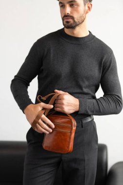 Koyu gri takım yakalı şık bir adam konyak deri bir çanta ile minimalist beyaz stüdyo arka planına karşı poz veriyor.