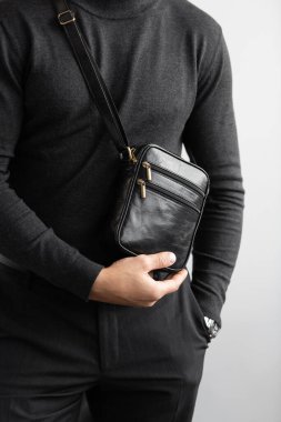 Kömür örgü yakalı bir erkek model ve beyaz bir stüdyo arka planına karşı siyah deri, dikey bir kurye çantası giyen terzi pantolonu.