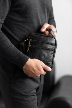 Kömür örgü yakalı bir erkek model ve beyaz bir stüdyo arka planına karşı siyah deri, dikey bir kurye çantası giyen terzi pantolonu.