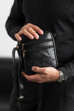 Kömür örgü yakalı bir erkek model ve beyaz bir stüdyo arka planına karşı siyah deri, dikey bir kurye çantası giyen terzi pantolonu.