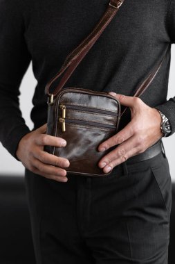 Mangal kömürü örgü ve pantolon giyen şık bir adam beyaz bir stüdyo arka planına karşı koyu kahverengi, dikey deri bir kurye çantası tutuyor..