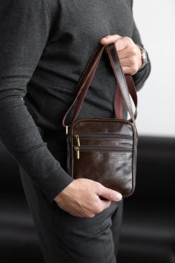 Mangal kömürü örgü ve pantolon giyen şık bir adam beyaz bir stüdyo arka planına karşı koyu kahverengi, dikey deri bir kurye çantası tutuyor..