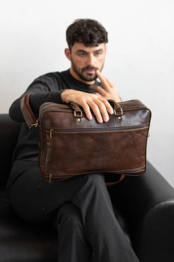 Kömür örgü ve terzi pantolonu giymiş profesyonel bir erkek model siyah deri bir koltukta oturuyor ve elinde özel kahverengi bir evrak çantası var.