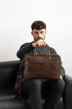 Kömür örgü ve terzi pantolonu giymiş profesyonel bir erkek model siyah deri bir koltukta oturuyor ve elinde özel kahverengi bir evrak çantası var.