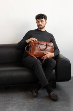 Kömür örgü ve terzi pantolonu giymiş profesyonel bir erkek model siyah deri bir koltukta oturuyor ve elinde özel kahverengi bir evrak çantası var.
