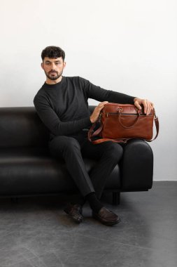 Kömür örgü ve terzi pantolonu giymiş profesyonel bir erkek model siyah deri bir koltukta oturuyor ve elinde özel kahverengi bir evrak çantası var.