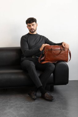 Kömür örgü ve terzi pantolonu giymiş profesyonel bir erkek model siyah deri bir koltukta oturuyor ve elinde özel kahverengi bir evrak çantası var.