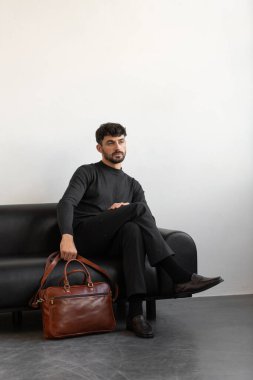 Kömür örgü ve terzi pantolonu giymiş profesyonel bir erkek model siyah deri bir koltukta oturuyor ve elinde özel kahverengi bir evrak çantası var.