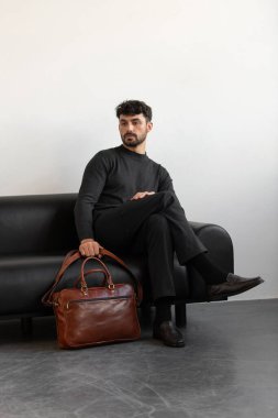 Kömür örgü ve terzi pantolonu giymiş profesyonel bir erkek model siyah deri bir koltukta oturuyor ve elinde özel kahverengi bir evrak çantası var.