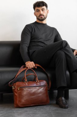 Kömür örgü ve terzi pantolonu giymiş profesyonel bir erkek model siyah deri bir koltukta oturuyor ve elinde özel kahverengi bir evrak çantası var.