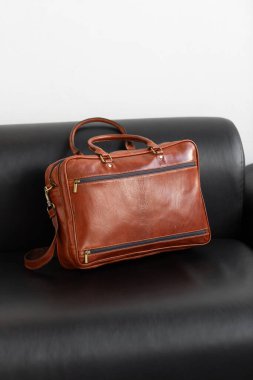 Profesyonel bir konyak çantası, çift kulplu ve ayarlanabilir kayışlı. Minimalist bir stüdyoda parlak siyah deri koltukta oturuyor.