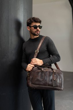 Güneş gözlüklü ve kömür örgülü kendinden emin bir adam koyu kahverengi bir evrak çantası tutarken modern minimalist bir iç mekanda dokulu bir sütuna yaslanıyor.