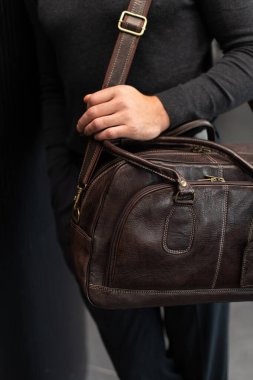 Kömür örgülü, kendine güvenen bir adam, modern, minimalist bir iç mekanda dokulu bir sütuna yaslanırken kahverengi deri bir evrak çantası tutuyor.