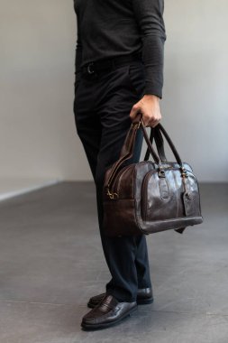 Kömür örgülü, kendine güvenen bir adam, modern, minimalist bir iç mekanda dokulu bir sütuna yaslanırken kahverengi deri bir evrak çantası tutuyor.