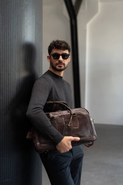 Güneş gözlüklü ve kömür örgülü kendinden emin bir adam koyu kahverengi bir evrak çantası tutarken modern minimalist bir iç mekanda dokulu bir sütuna yaslanıyor.