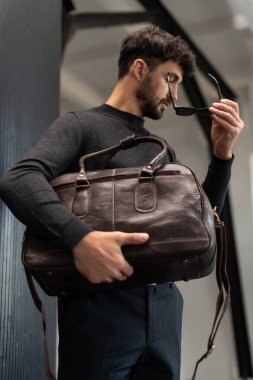 Kömür örgülü, kendine güvenen bir adam, modern, minimalist bir iç mekanda dokulu bir sütuna yaslanırken kahverengi deri bir evrak çantası tutuyor.