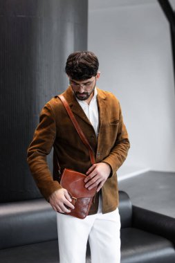 Kahverengi kadife ceketli, beyaz gömlekli şık bir adamın portresi. Stüdyoda bir konyak deri bir kurye çantası tutuyor.
