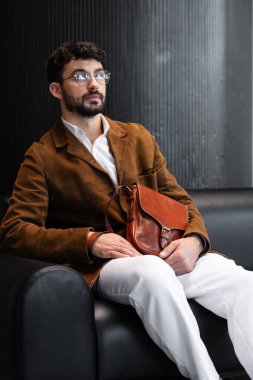 Kahverengi kadife ceketli ve gözlüklü sofistike bir adam siyah deri koltukta oturmuş minimalist bir stüdyoda, deri bir çanta taşıyor.