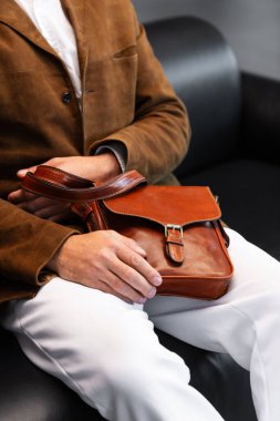 Kahverengi kadife ceketli bir adamın minimalist bir stüdyoda otururken tuttuğu metal tokalı birinci sınıf bir konyak postacı çantasının yakın çekimi...