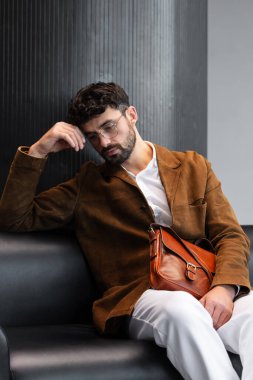 Kahverengi kadife ceketli ve gözlüklü sofistike bir adam siyah deri koltukta oturmuş minimalist bir stüdyoda, deri bir çanta taşıyor.