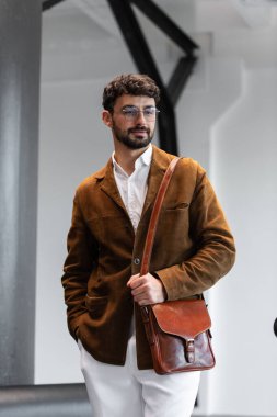 Kahverengi kadife ceketli, beyaz gömlekli şık bir adamın portresi. Stüdyoda bir konyak deri bir kurye çantası tutuyor.