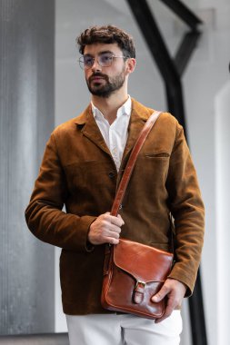 Kahverengi kadife ceket ve beyaz gömlekli şık bir adamın portresi. Minimalist bir stüdyoda, deri bir kurye çantası tutuyor.