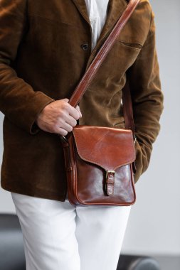 Minimalist bir stüdyoda kahverengi kadife ceketli bir adamın tuttuğu metal tokalı birinci sınıf bir konyak postacı çantasının yakın çekimi...