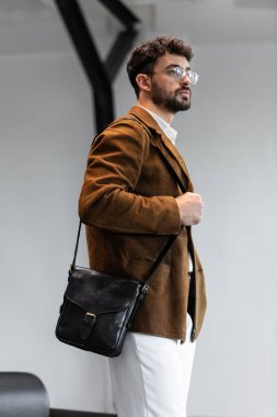 Kahverengi kadife ceketli ve beyaz gömlekli şık bir adamın portresi. Minimalist bir stüdyoda siyah deri bir kurye çantası tutuyor.