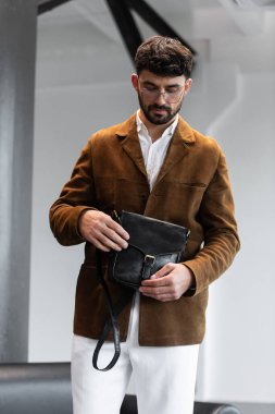 Kahverengi kadife ceketli ve beyaz gömlekli şık bir adamın portresi. Minimalist bir stüdyoda siyah deri bir kurye çantası tutuyor.