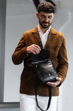 Kahverengi kadife ceketli ve beyaz gömlekli şık bir adamın portresi. Minimalist bir stüdyoda siyah deri bir kurye çantası tutuyor.