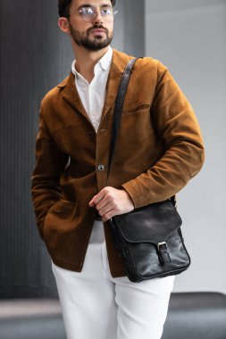 Kahverengi kadife ceketli ve beyaz gömlekli şık bir adamın portresi. Minimalist bir stüdyoda siyah deri bir kurye çantası tutuyor.