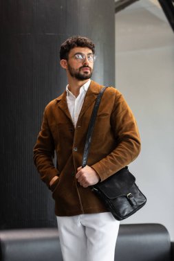 Kahverengi kadife ceketli ve beyaz gömlekli şık bir adamın portresi. Minimalist bir stüdyoda siyah deri bir kurye çantası tutuyor.