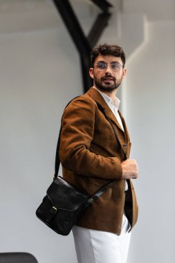 Kahverengi kadife ceketli ve beyaz gömlekli şık bir adamın portresi. Minimalist bir stüdyoda siyah deri bir kurye çantası tutuyor.