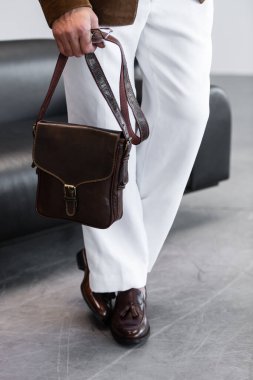 Beyaz pantolonlu ve kahverengi deri ayakkabılı bir adamın, minimalist bir stüdyoda koyu kahverengi deri bir kurye çantası taşıdığı görüntüler..