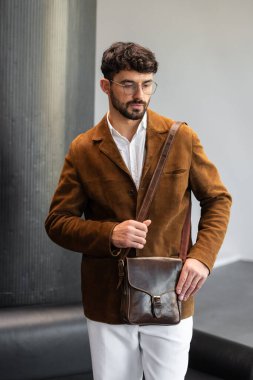 Kahverengi kadife ceket ve beyaz gömlekli şık bir adamın portresi. Minimalist bir stüdyoda, deri bir kurye çantası tutuyor.