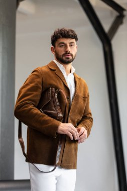 Kahverengi kadife ceket ve beyaz gömlekli şık bir adamın portresi. Minimalist bir stüdyoda, deri bir kurye çantası tutuyor.