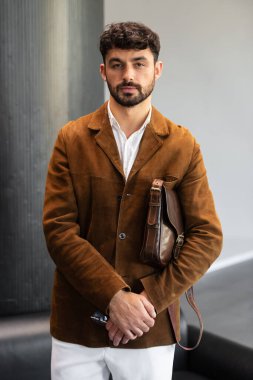 Kahverengi kadife ceket ve beyaz gömlekli şık bir adamın portresi. Minimalist bir stüdyoda, deri bir kurye çantası tutuyor.