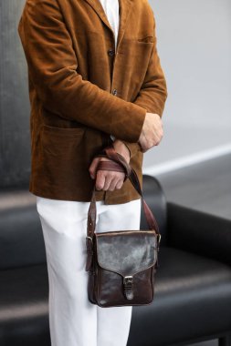 Kahverengi kadife ceketli bir adamın, minimalist bir stüdyoda kemer takılı kahverengi deri bir kurye çantası taşıdığı yakın plan.