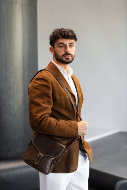 Kahverengi kadife ceket ve beyaz gömlekli şık bir adamın portresi. Minimalist bir stüdyoda, deri bir kurye çantası tutuyor.