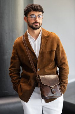 Kahverengi kadife ceketli ve beyaz gömlekli şık bir adamın portresi. Minimalist bir stüdyoda siyah deri bir kurye çantası tutuyor.