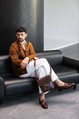 Kahverengi kadife ceketli ve beyaz pantolonlu şık bir adam siyah deri koltukta oturuyor ve minimalist bir stüdyoda kahverengi deri bir kurye çantası taşıyor..