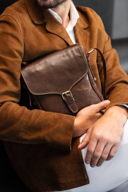 Minimalist bir stüdyoda oturan kahverengi kadife ceketli bir adamın tuttuğu metal tokalı özel deri bir kurye çantasının yakın çekimi....