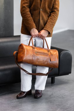 Süet ceket ve beyaz pantolon giyen kahverengi spor çantalı bir adamın yakın plan fotoğrafı..
